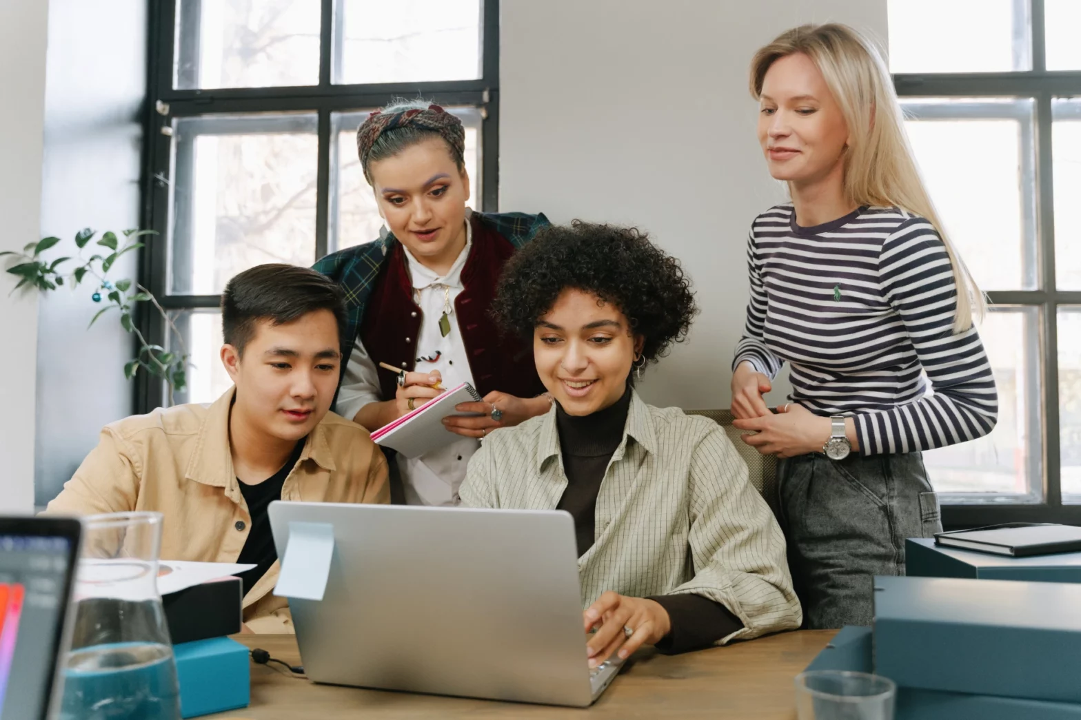 Four colleagues gathered around a desk, looking closely at a laptop screen together during a collaborative team meeting