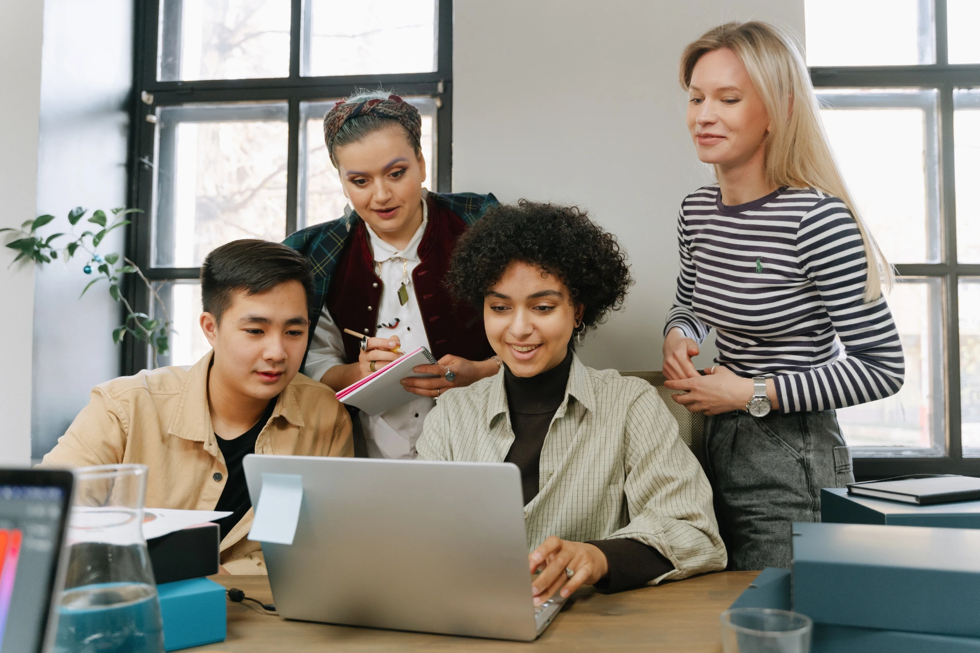 Four colleagues gathered around a desk, looking closely at a laptop screen together during a collaborative team meeting