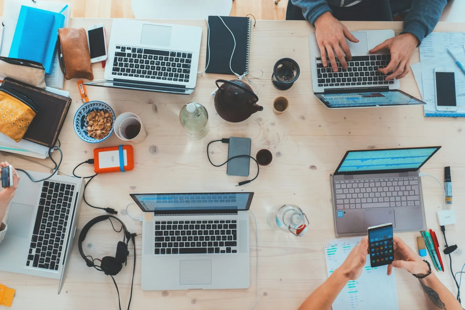 Overhead view of a busy desk with five laptops, phones, notebooks, and various office supplies