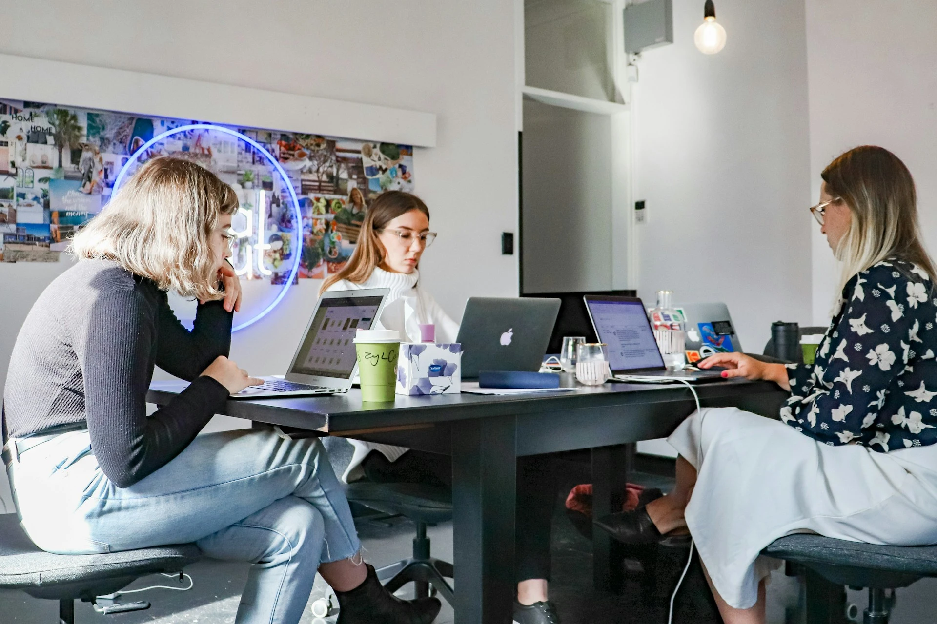 Three women sit at a work table, each focused on their laptops. The atmosphere is focused and productive.