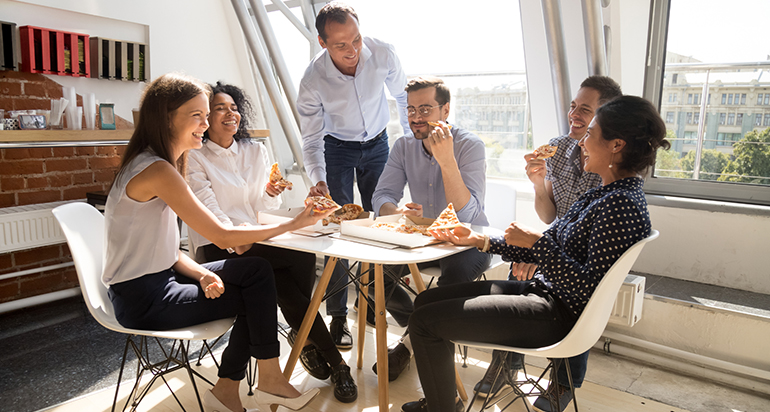 Employees eating pizza together at a table in a brightly lit room during their lunch hour