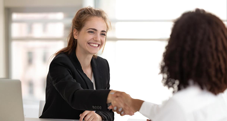 A young smiling woman in a black blazer shaking hands with an HR representative during onboarding