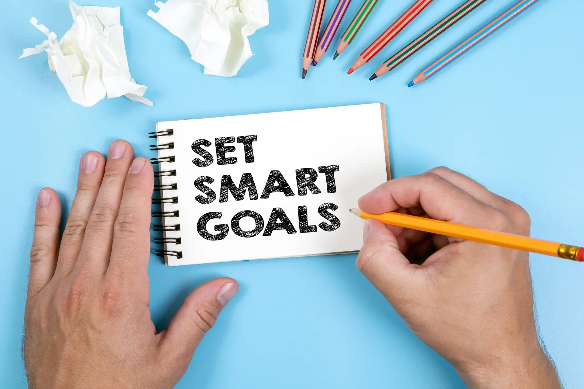 An individual is inscribing in bold letters on a notepad with a pencil the phrase “SET SMART GOALS.” 