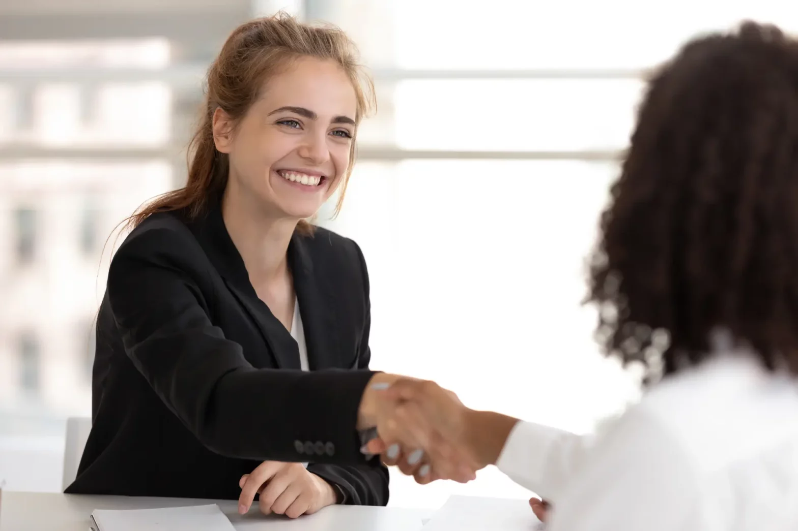 A young smiling woman in a black blazer shaking hands with an HR representative during onboarding
