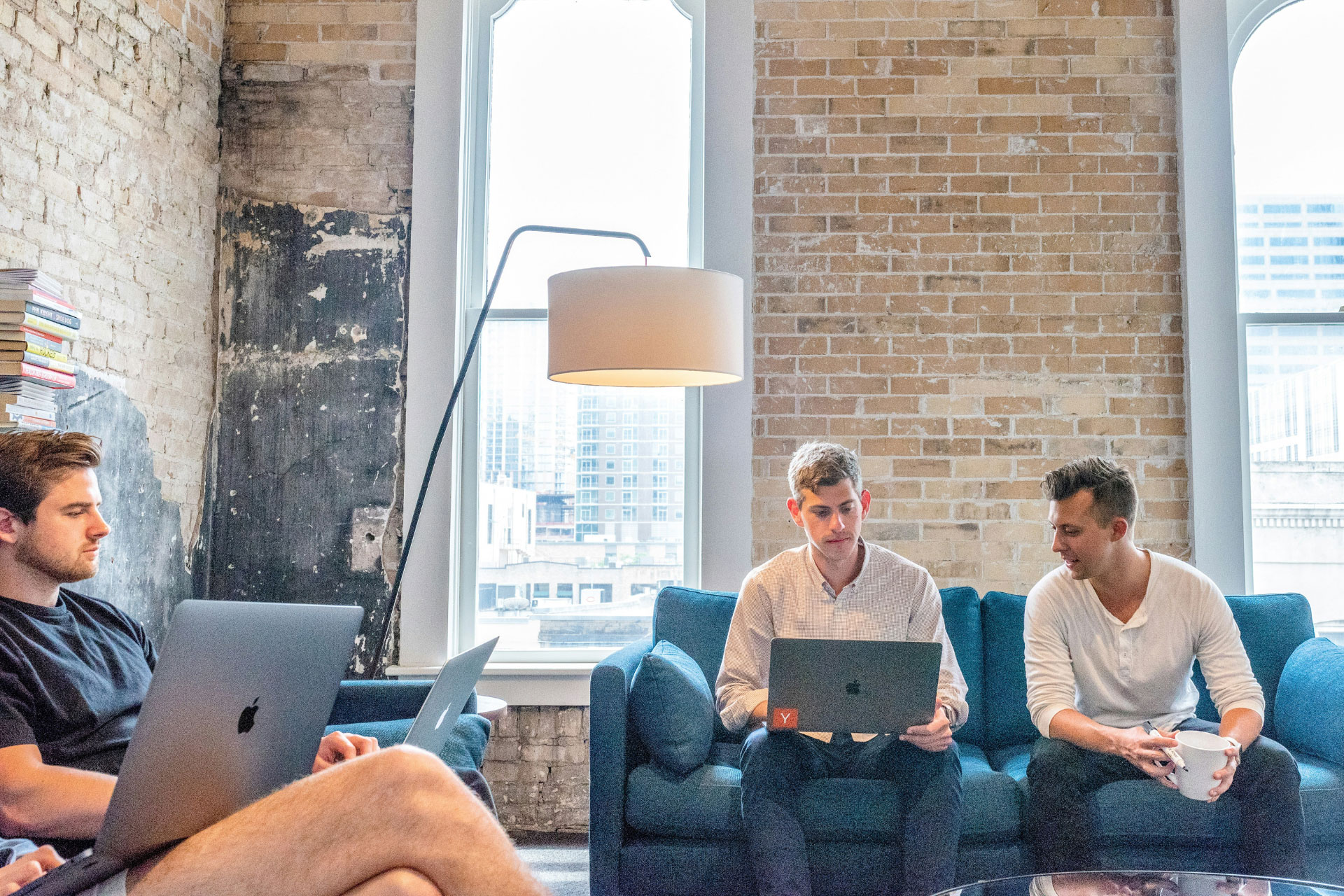 A few men sitting on couches working on their laptops in an office with brick walls