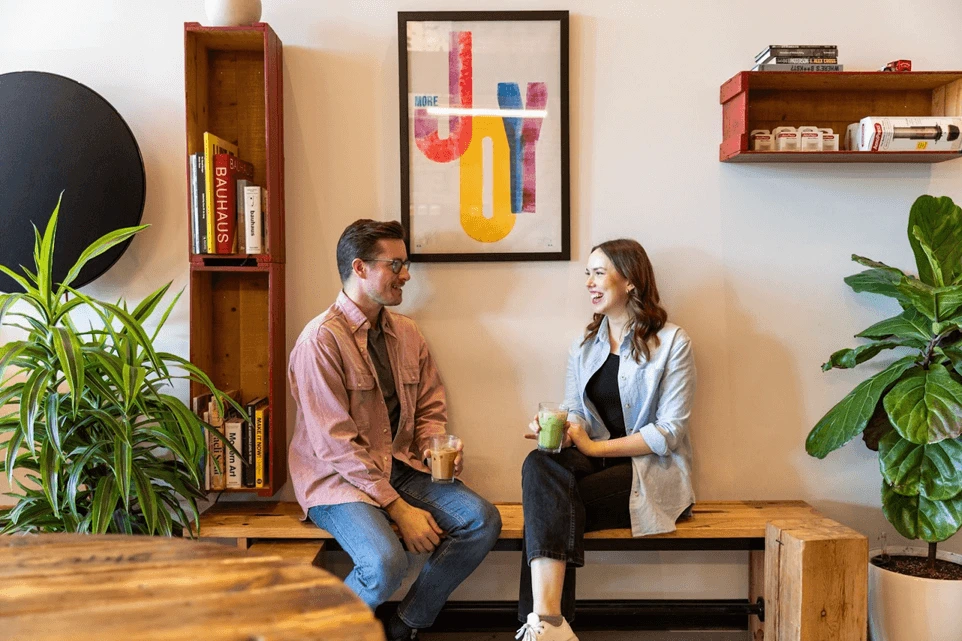 Two employees sit side by side on a wooden bench in an office, each holding a smoothie.