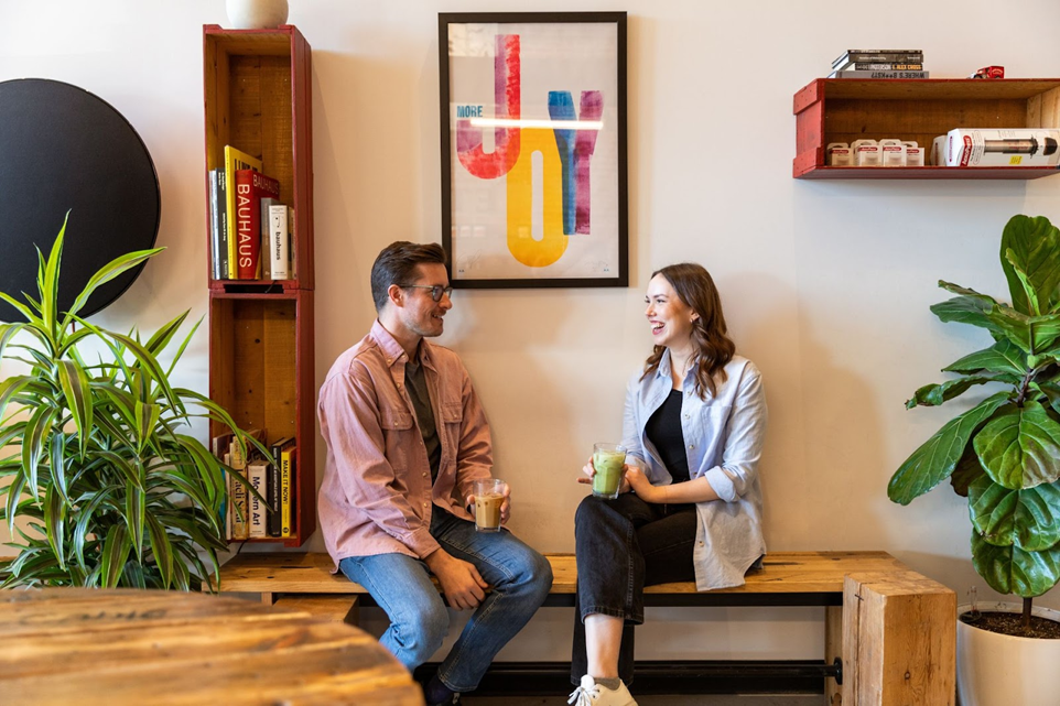 Two employees sit side by side on a wooden bench in an office, each holding a smoothie.