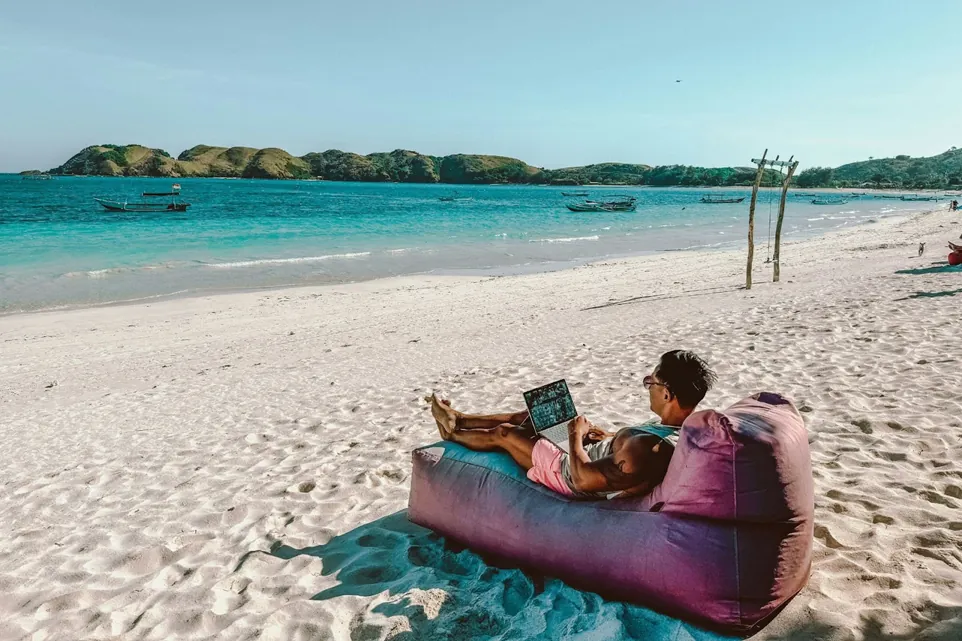 An employee sits on a bean bag on a beach, while working on his laptop.