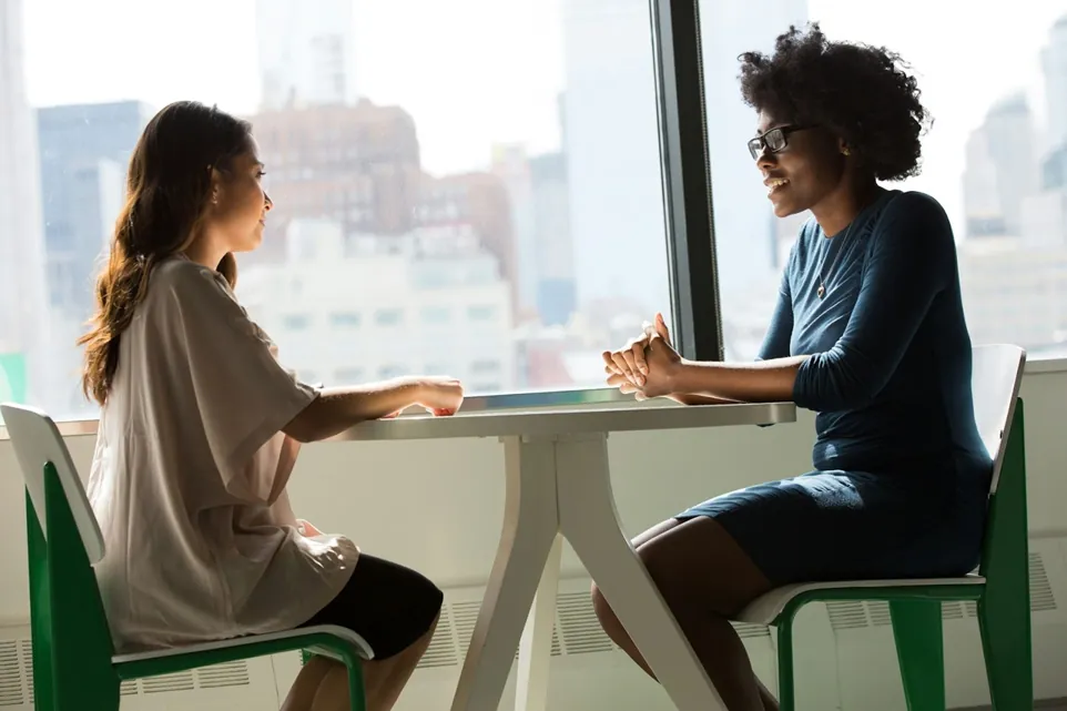 Two young women sit face to face at a circular table for an interview. 