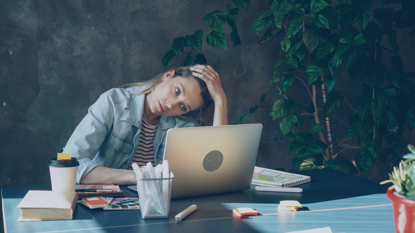 A young woman sits at her desk with a laptop, her head tilted to the side, appearing visibly stressed