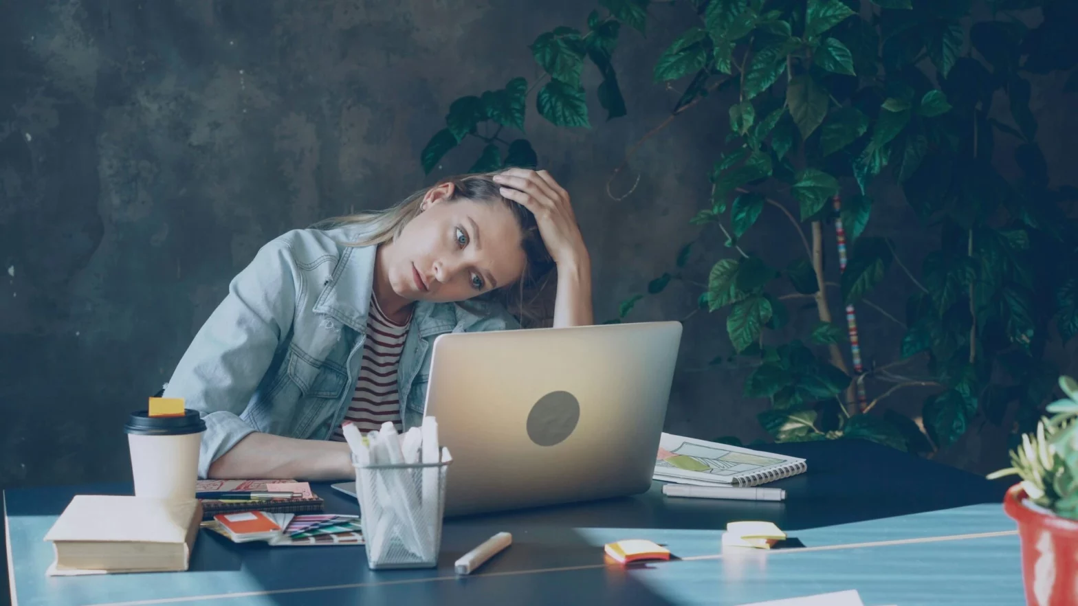 A young woman sits at her desk with a laptop, her head tilted to the side, appearing visibly stressed