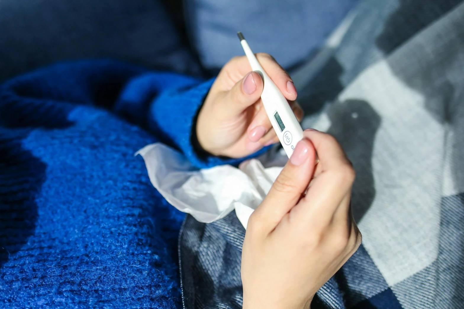Someone holding a thermometer underneath a blanket with a tissue, wondering when to request a sick note